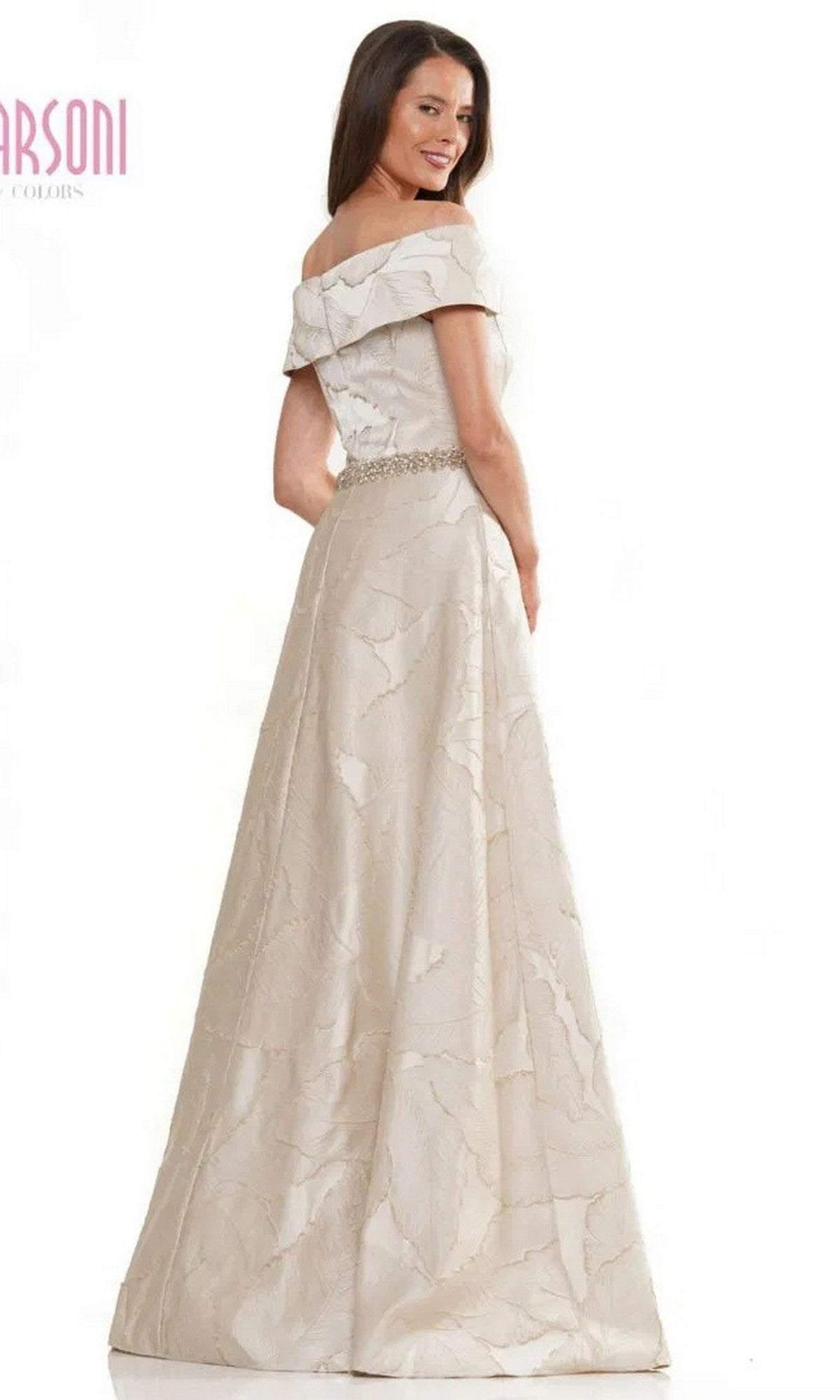 A woman wearing the Marsoni MV1265 long formal dress, featuring an off-the-shoulder design, subtle leaf pattern, and beaded waistline, stands smiling at the camera.